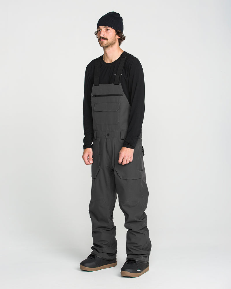 A man wearing a black beanie, a black long-sleeve shirt, and dark gray Volcom Men’s Roan Bib overalls stands facing slightly left against a plain white background. The stylish Volcom Men’s Roan Bib is reminiscent of recycled snow pants, seamlessly blending fashion with function.