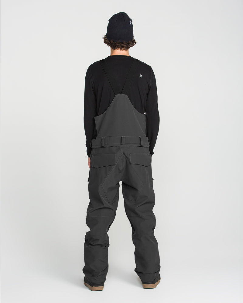 A person stands facing away, wearing a black beanie, a black long-sleeve shirt, and the Volcom Men’s Roan Bib in dark gray—featuring multiple pockets and snow pant styling—against a plain white background.