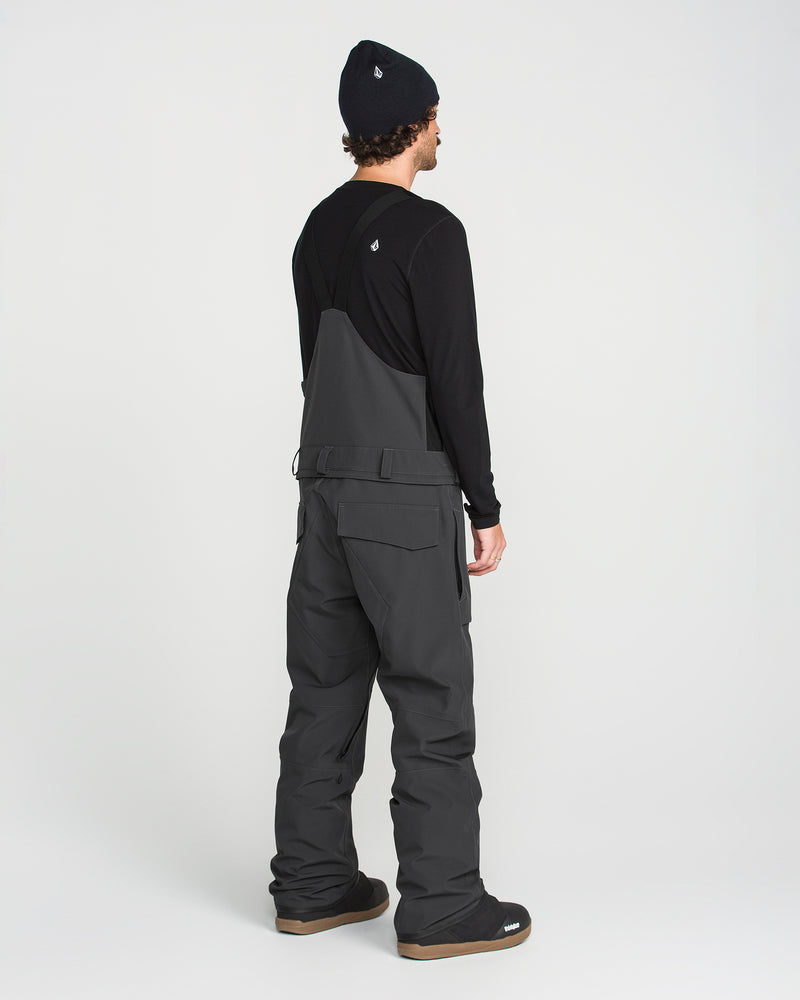 A person stands facing away, wearing a dark beanie, a black long-sleeve shirt, and the Volcom Men’s Roan Bib in gray over boots, set against a plain white background.