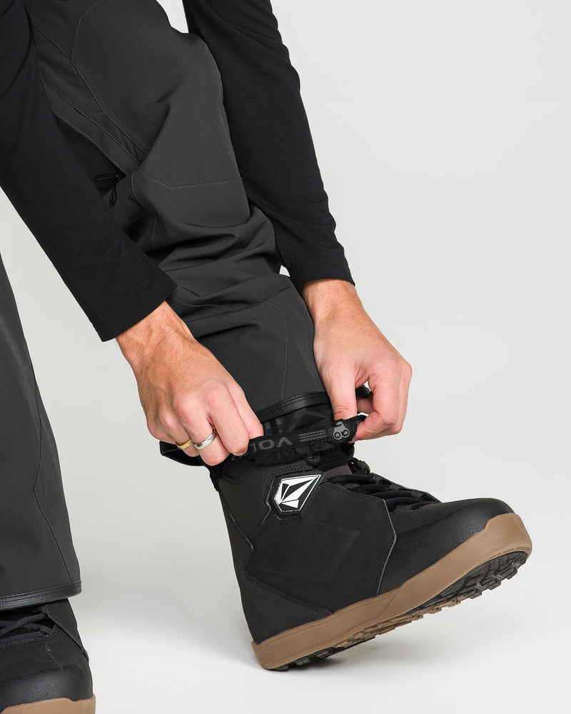 A person wearing Volcom Men’s Roan Bib fastens the strap on a black snowboard boot with a brown sole. The background is light and uncluttered.