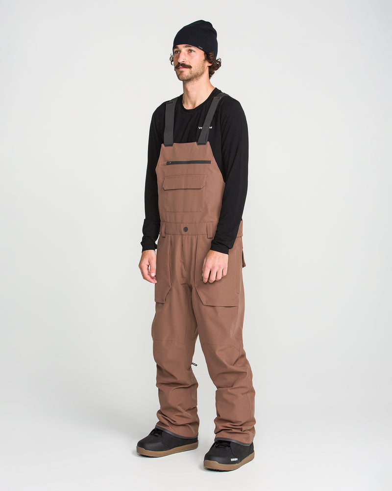 A man with a mustache stands facing forward against a plain white background, wearing a black beanie, a black long-sleeve shirt, and brown Men’s Roan Bib overalls by Volcom, crafted from recycled stretch Oxford. He is also wearing black shoes.