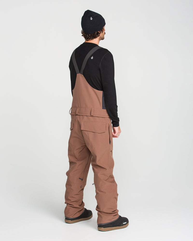 A person stands facing away against a plain white background, wearing a black long-sleeve shirt, Volcom Men’s Roan Bib overall snow pants in brown crafted from recycled stretch Oxford for durability and comfort, black boots, and a black beanie.