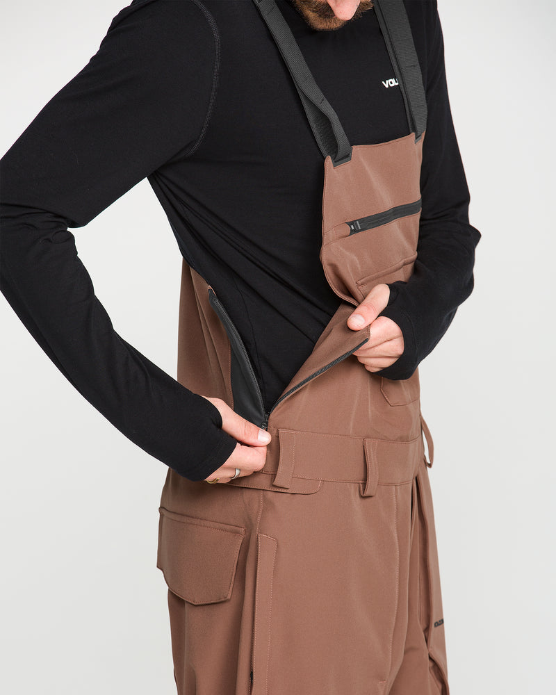A person wearing Volcom Men’s Roan Bib overalls in brown, crafted from recycled stretch Oxford fabric, and a black long-sleeve shirt is unzipping the side of the overalls. The image emphasizes the clothing and hand, with the head partially out of frame.