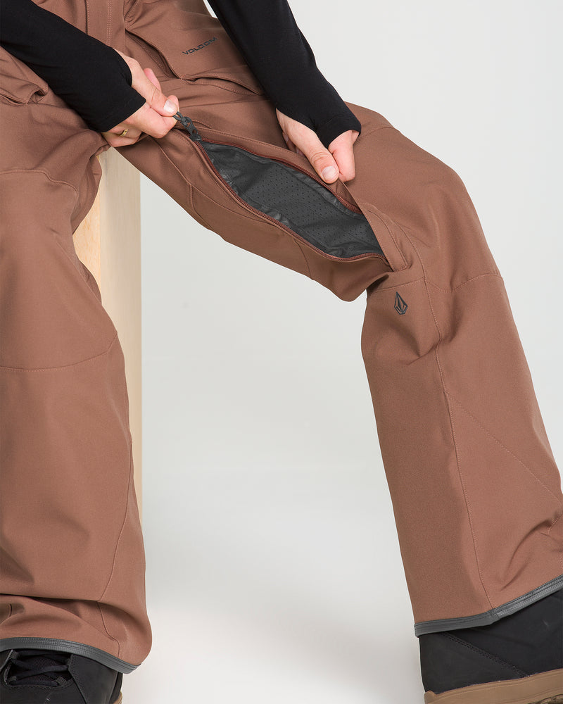 A person wearing Volcom Men’s Roan Bib pants in brown sits on a light wooden stool, unzipping the ventilation panel on the side of their recycled stretch Oxford pants. Only their lower body and hands are visible.