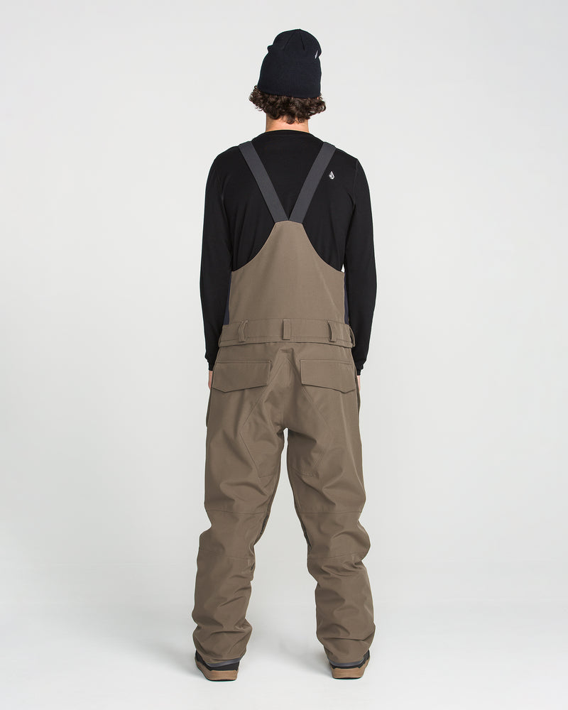 A person stands facing away, wearing a black long-sleeve shirt, a black beanie, boots, and the Volcom Men’s Roan Bib Overall in brown—crafted from recycled stretch Oxford fabric for comfort and durability with adjustable suspenders—against a white background.