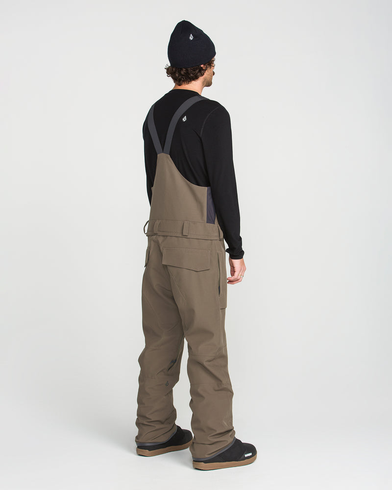 A person stands facing away on a plain white background, dressed in a black beanie, black long-sleeve shirt, and the Volcom Men’s Roan Bib Overall in khaki. Made from recycled stretch Oxford fabric, this bib showcases 15K protection.