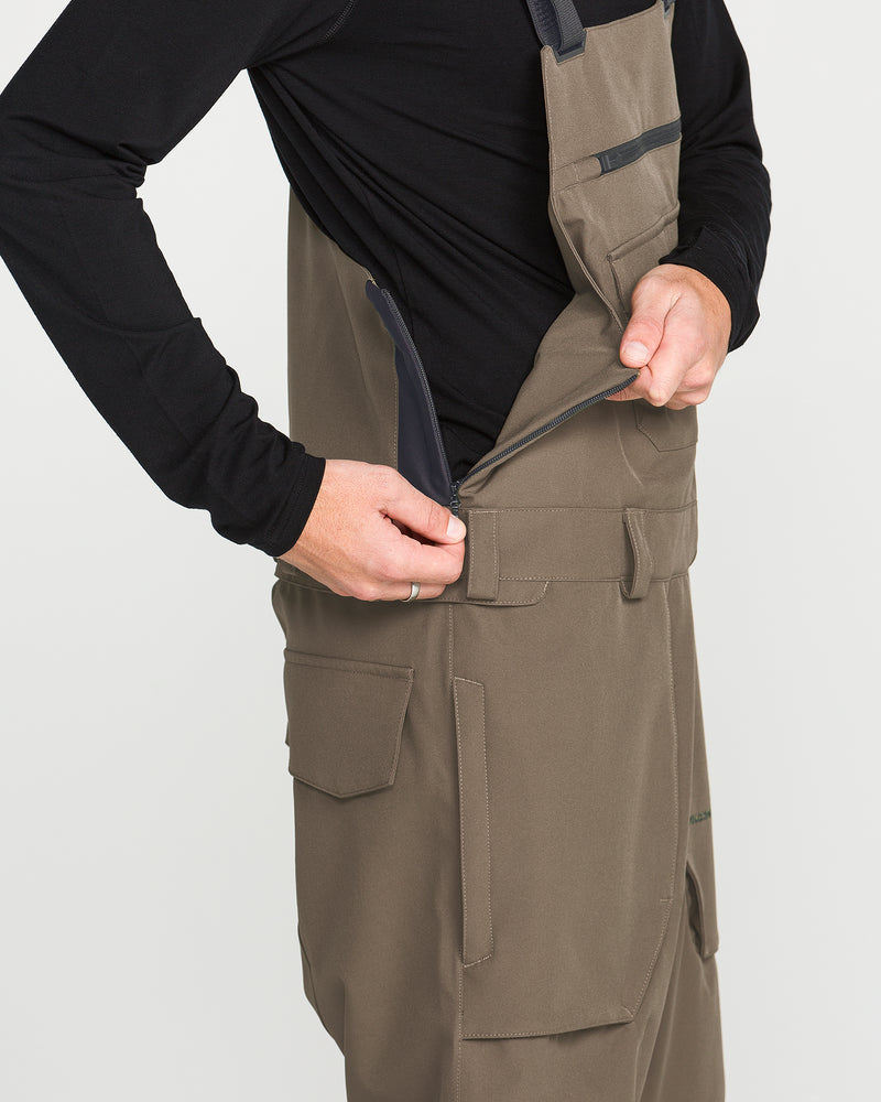 A person wearing the Volcom Men’s Roan Bib, crafted from recycled stretch Oxford fabric and paired with a black shirt, demonstrates unzipping a side zipper on the bib to reveal their shirt underneath while highlighting the bib’s 15K protection.