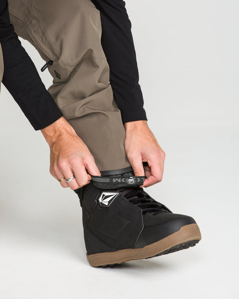 A person wearing Volcom Men’s Roan Bib Overalls in khaki, crafted from recycled stretch Oxford fabric, and a black long-sleeve shirt tightens the strap on a black snowboarding boot with a brown sole, ready to take on the slopes with 15K protection.