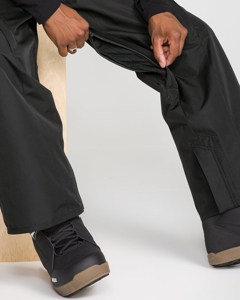 A person is sitting and unzipping a side vent on black Volcom Men’s Kleveland Pants, wearing black shoes with brown soles, against a plain background.
