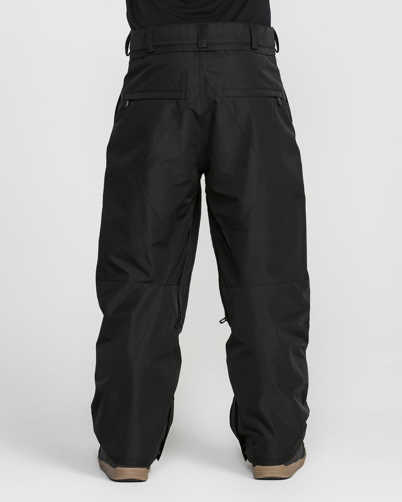 A person is shown from the back wearing loose-fitting black Volcom Men’s Kleveland Pants, with visible pockets and belt loops; these snow pants, featuring V-Science 2-Layer construction, cover brown shoes against a plain white background.