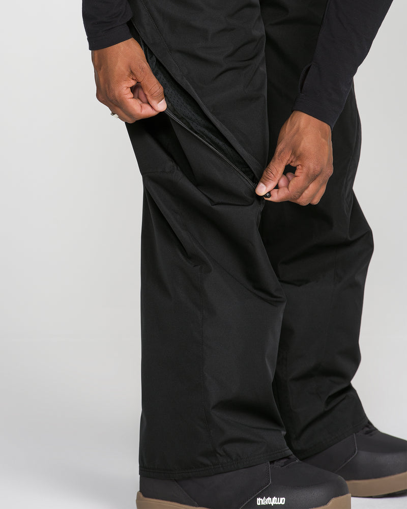 A person unzips the side of black Volcom Men’s Kleveland Pants, revealing a mesh lining characteristic of this snowboard pant, while wearing black shoes with brown soles.