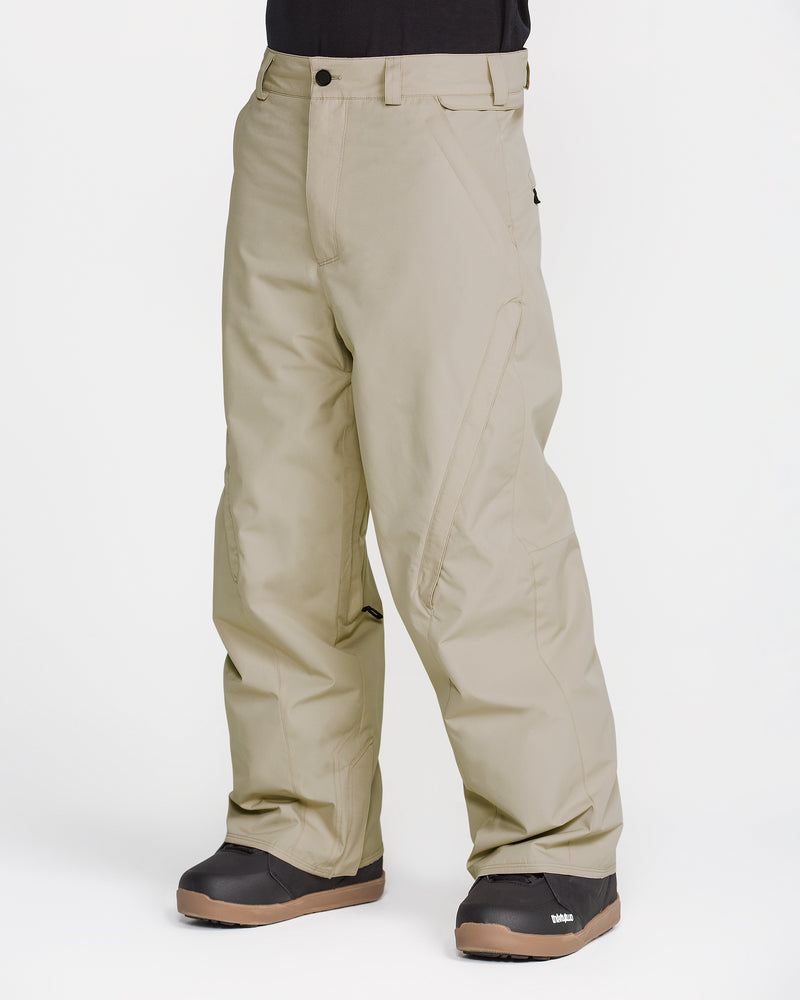 A person is wearing the Volcom Men’s Kleveland Pants in a loose, baggy beige fit, paired with black winter boots, standing against a plain white background.