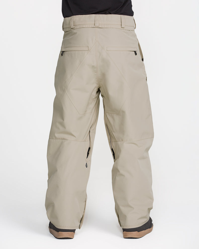 A person is shown from the waist down, wearing Volcom Men’s Kleveland Pants in beige. The loose-fitting snowboard pants feature back pockets and zipper details, set against a plain white background.