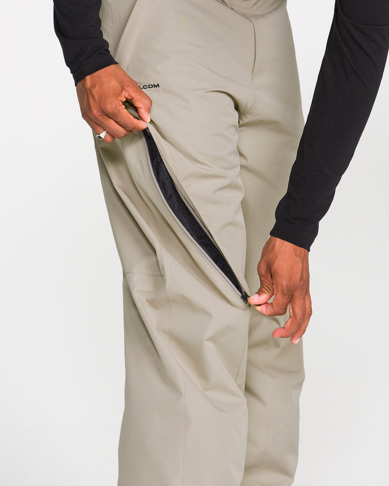 Wearing a black long-sleeve shirt, a person unzips the black-zippered side pocket on a pair of beige Volcom Men’s Kleveland Pants.