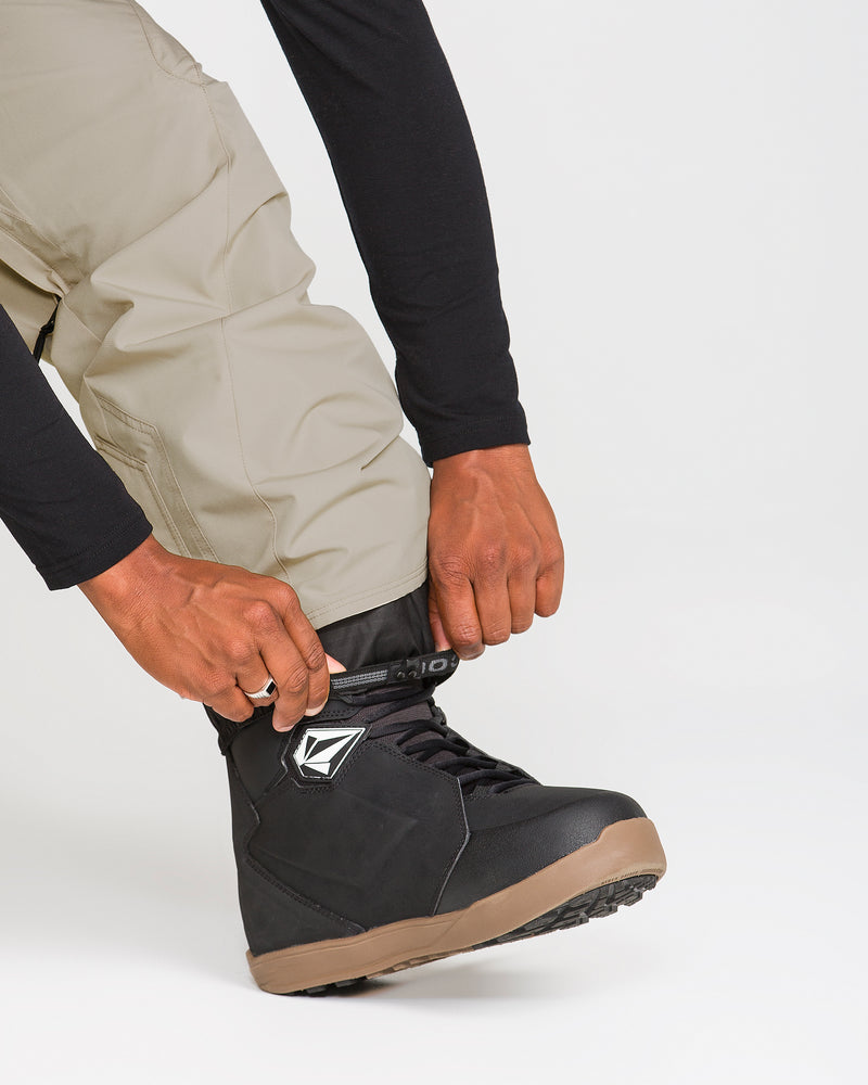Wearing Volcom Men’s Kleveland Pants in a baggy fit and a black long-sleeve shirt, a person secures the strap on a black snowboarding boot with a tan sole on their right foot.