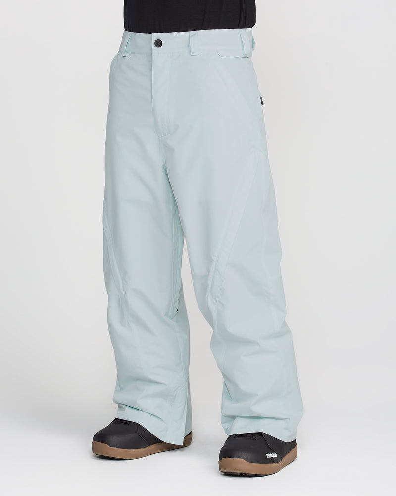 A person is shown from the waist down, wearing Volcom Men’s Kleveland Pants in a light blue, baggy fit along with black snow boots, standing against a plain white background. The image highlights the V-Science 2-Layer construction of these snowboard pants for superior weather protection and comfort.