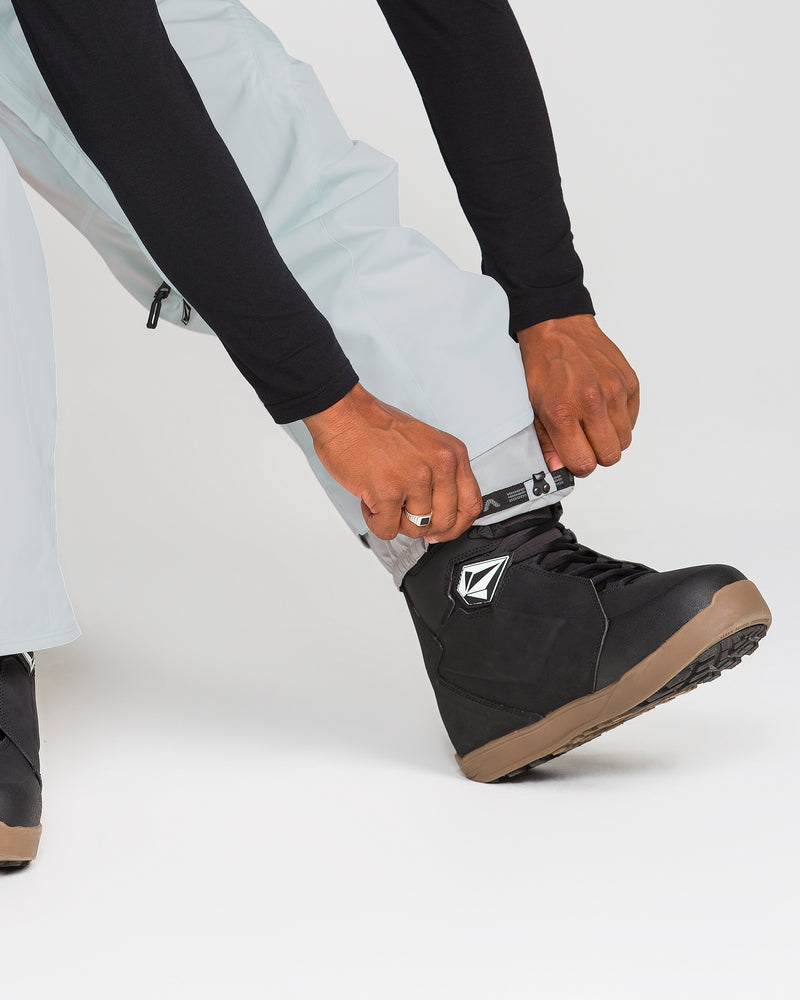 A person adjusts the cuff of Volcom Men’s Kleveland Pants in a light color over a black snowboarding boot with a brown sole and a geometric logo. The background is plain white.