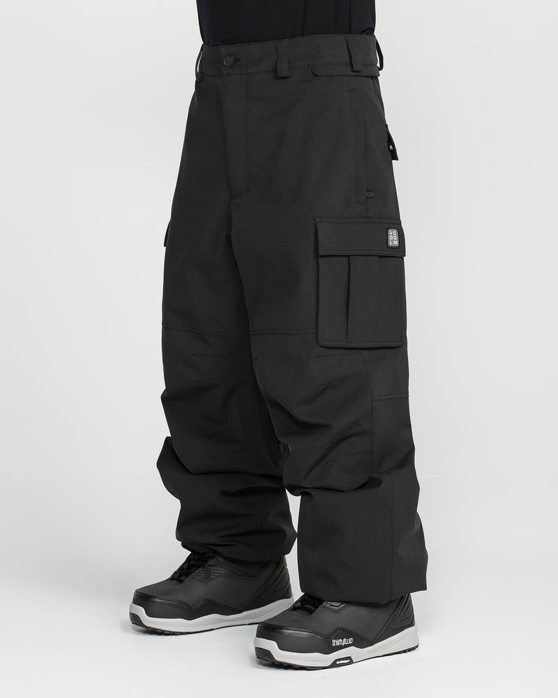 A person is wearing the Volcom Men’s Nwrk 20K Baggy Pants—loose-fitting black cargo pants with multiple pockets, made from recycled shell fabric—paired with black and gray athletic shoes, standing against a plain white background. The relaxed, baggy design of the pants provides a comfortable fit and drapes over part of the shoes.
