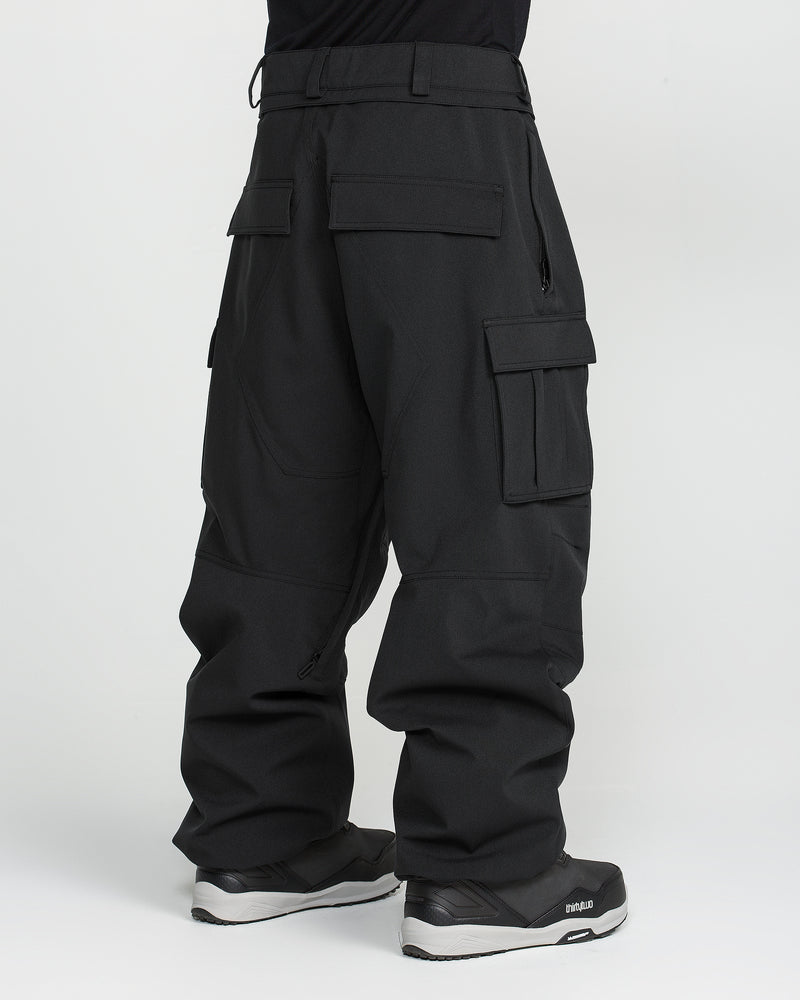 A person wearing the Volcom Men’s Nwrk 20K Baggy Pants—loose-fitting black cargo pants crafted from recycled shell fabric with multiple large pockets—paired with black sneakers, stands against a plain white background.