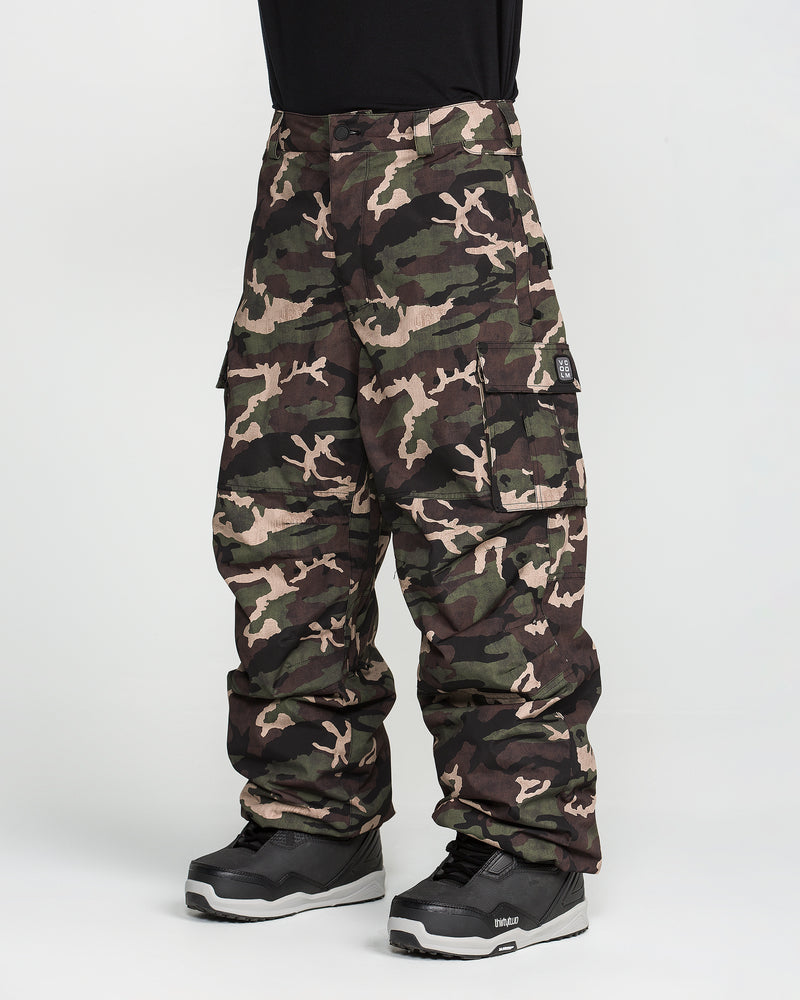 A person wearing Volcom Men’s Nwrk 20K Baggy Pants in a camouflage cargo style and black snow boots, standing against a white background; only the lower half of their body is visible.