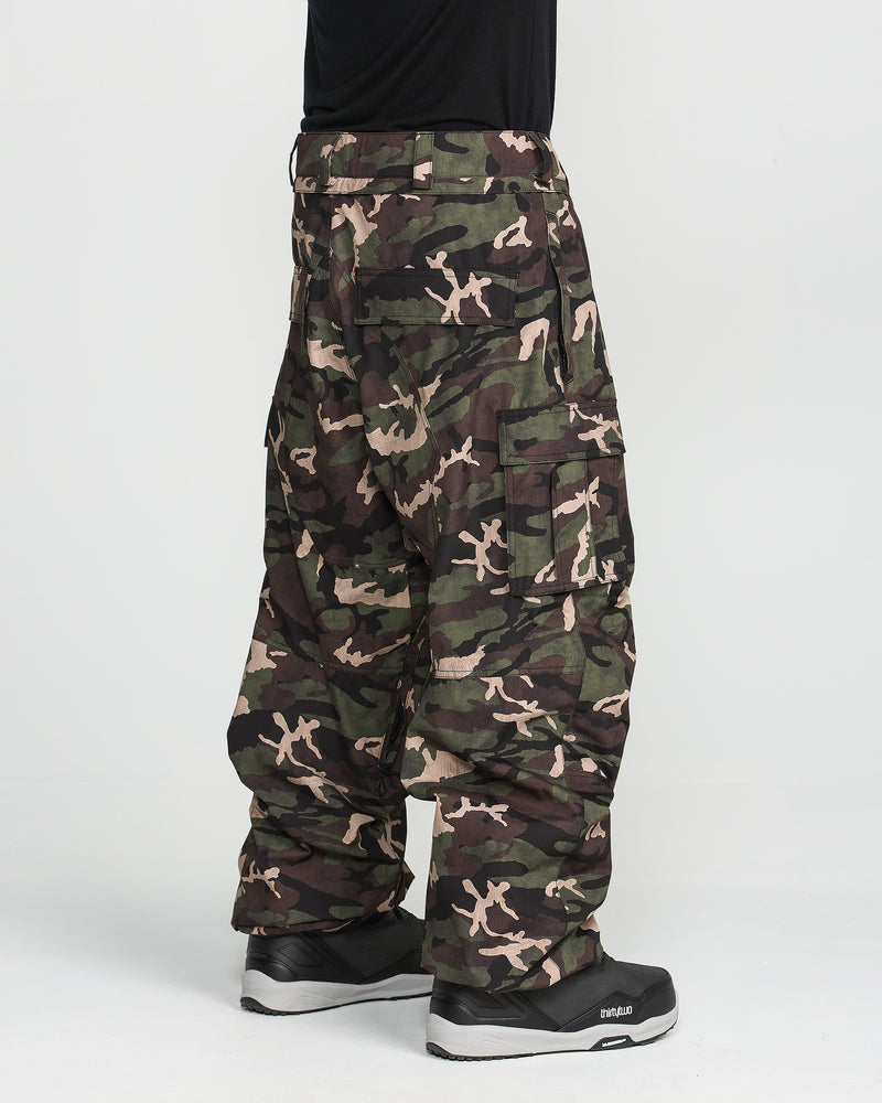 A person wearing Volcom Men’s Nwrk 20K Baggy Pants styled like snowboard pants, paired with black sneakers, stands against a plain white background with only the lower half of the body visible.