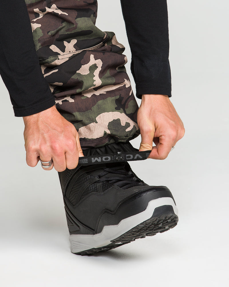A person wearing Volcom Men’s Nwrk 20K Baggy Pants adjusts the strap on a black snowboarding boot, preparing for use. Only their hands, lower legs clad in the pants, and part of the boot are visible against a plain background.