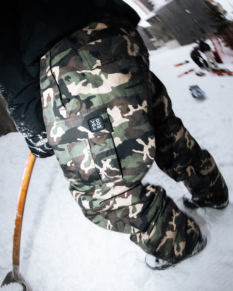 A person wearing Volcom Men’s Nwrk 20K Baggy Pants with a breathable lining stands on snow, holding a wooden-handled tool. The pants’ pocket features a visible ROC 200 patch. Nearby objects and snowy scenery appear in the background, captured with a fisheye lens effect.
