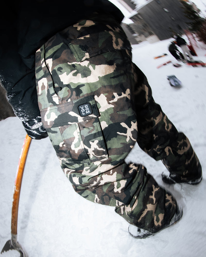 A person wearing Volcom Men’s Nwrk 20K Baggy Pants with a breathable lining stands on snow, holding a wooden-handled tool. The pants’ pocket features a visible ROC 200 patch. Nearby objects and snowy scenery appear in the background, captured with a fisheye lens effect.