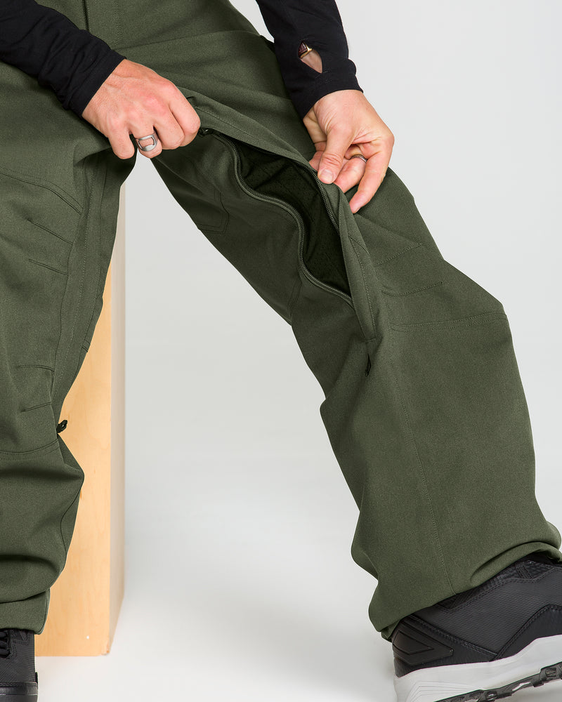 A person sits on a wooden bench, unzipping the side of olive green Volcom Men’s Nwrk 20K Baggy Pants to reveal the interior, with only their lower body and hands in view.