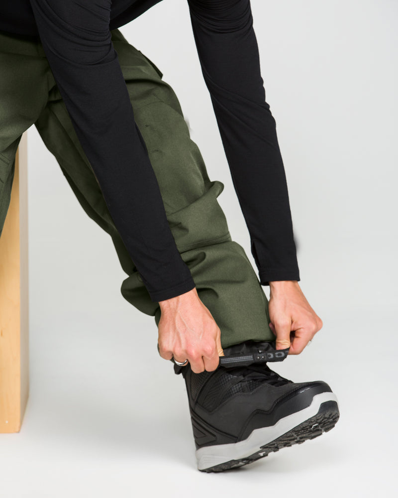 A person wearing Volcom Men’s Nwrk 20K Baggy Pants in green and a black long-sleeve shirt is tightening the strap on a black and gray winter boot. Only their lower body and hands are visible, drawing attention to the pants’ durable 20K waterproof fabric.