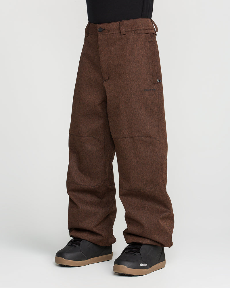 A person is wearing Volcom Men’s Snow Billow Pants in brown with an adjustable waist, paired with black snow boots, standing against a plain white background.