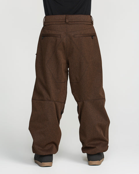 A person is seen from the back wearing Volcom Men’s Snow Billow Pants in brown, featuring an adjustable waist, paired with a black shirt and black shoes, and standing against a plain white background.