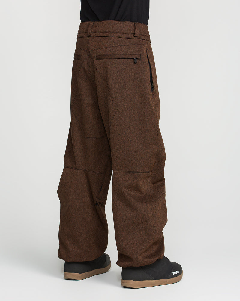 A person is shown from the waist down wearing Volcom Men’s Snow Billow Pants in brown with black boots, standing against a white background. The pants have zipper pockets, seam detailing at the knees, and an adjustable waist.