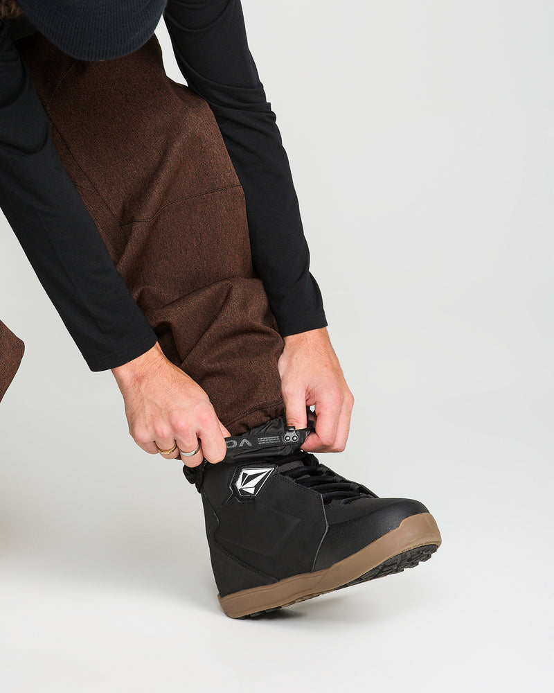 A person wearing Volcom Men’s Snow Billow Pants in brown, featuring an adjustable waist, and a black long-sleeve shirt adjusts the top of a black snow boot with a tan sole. The background is plain white.