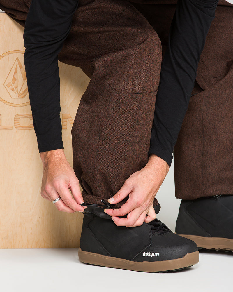Wearing Volcom Men’s Snow Billow Pants in brown, a person kneels before a wooden box with a faint logo, adjusting the adjustable cuff over a black winter boot with a tan sole.