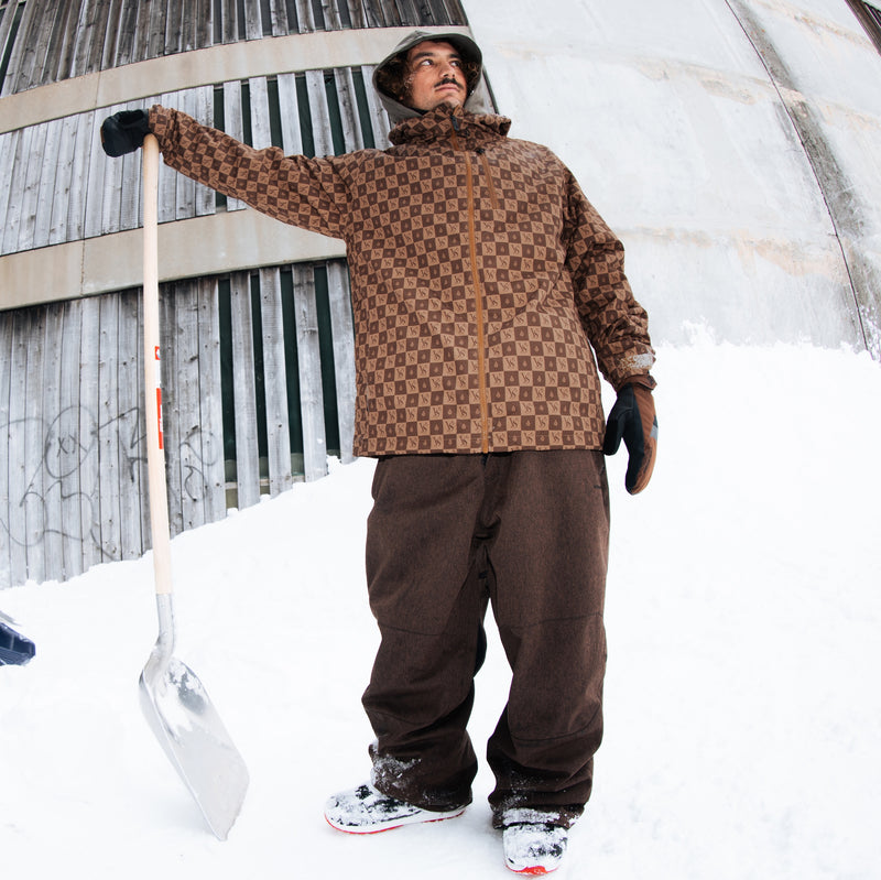 A man wearing a brown and tan checkered winter jacket, brown snow pants, and a hat stands in the snow holding a shovel. The background shows a snowy outdoor setting with a wooden fence and a curved concrete wall.