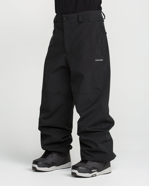 A person wearing loose-fitting black Volcom Men’s Snow Billow Pants, made with 10K waterproof V-Science 2-Layer fabric, and gray-black snow boots stands against a plain white background. Only the lower body and legs are visible; the upper body is cropped out.