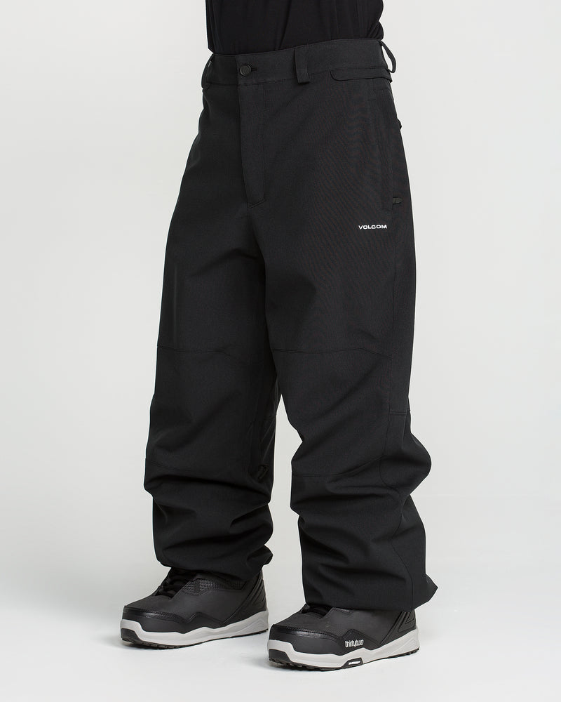 A person wearing loose-fitting black Volcom Men’s Snow Billow Pants, made with 10K waterproof V-Science 2-Layer fabric, and gray-black snow boots stands against a plain white background. Only the lower body and legs are visible; the upper body is cropped out.