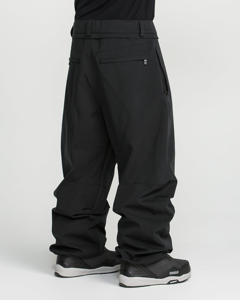 A person is shown standing sideways against a plain white background, wearing Volcom Men’s Snow Billow Pants in black and matching black sneakers. The upper body is out of frame. These men’s snow pants feature zippered pockets, reinforced stitching, and are crafted with V-Science 2-Layer fabric for reliable 10K waterproof protection.