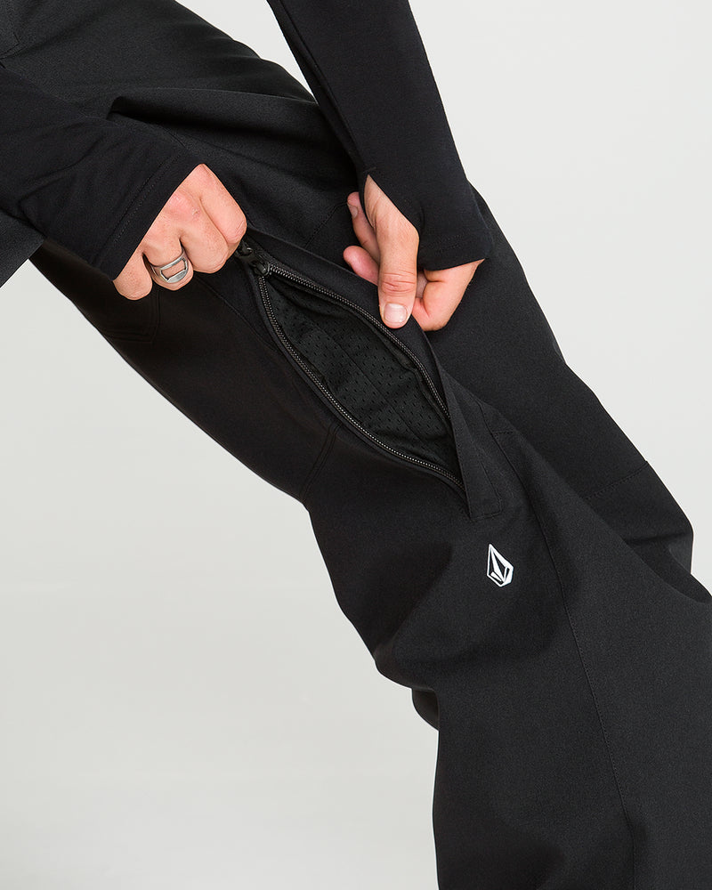 A person unzips a vent on the side of black Volcom Men’s Snow Billow Pants, revealing mesh fabric inside. Crafted with V-Science 2-Layer material and boasting 10K waterproof protection, the pants feature a small, white geometric logo near the knee.