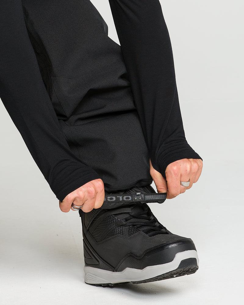 A person wearing black Volcom Men’s Snow Billow Pants and black shoes, likely featuring V-Science 2-Layer fabric and 10K waterproof protection, adjusts a strap at the ankle of their pants. Their hands are visible against a plain, light-colored background.
