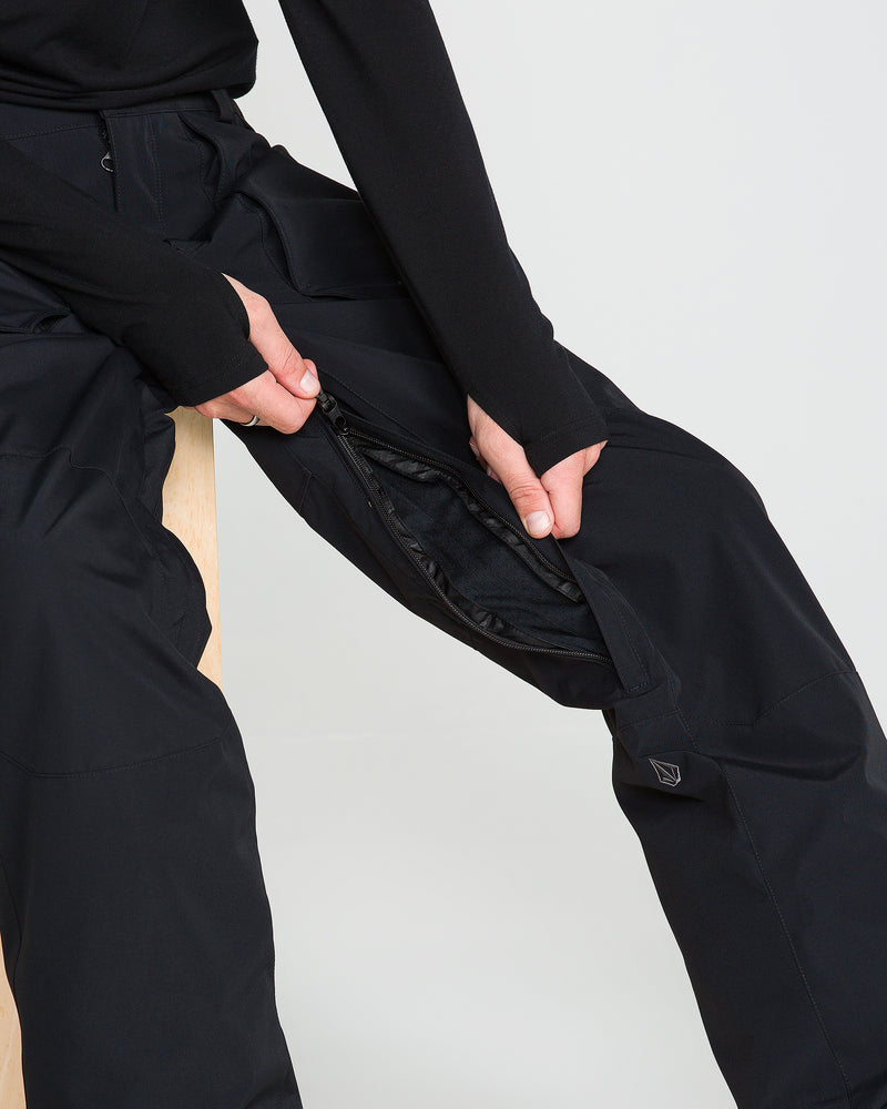A person seated, unzipping the side of Volcom Men’s Roan 20K Pants in black to reveal an inner layer. Only the lower body and hands are visible against a plain background.