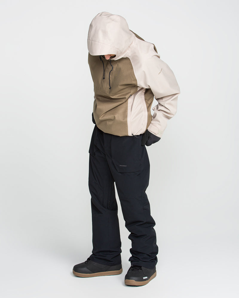 A person wearing a beige and brown hooded jacket, black pants, and dark shoes stands against a plain white background, looking down with hands in pockets. The outfit is completed with Volcom Men’s Roan 20K Pants—breathable ski pants crafted from recycled fabric snow pants—offering comfort and sustainability for outdoor adventures.