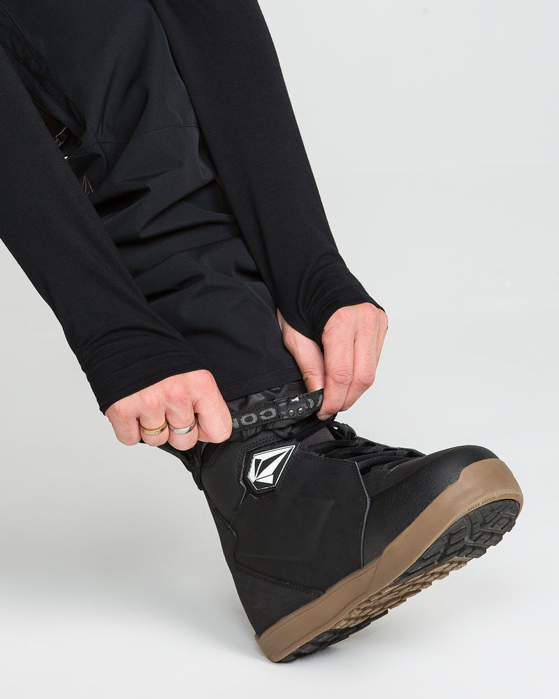 A person wearing Volcom Men’s Roan 20K Pants in black and a black shoe with a brown sole adjusts a patterned strap around their ankle. Only their hands and lower legs are visible.