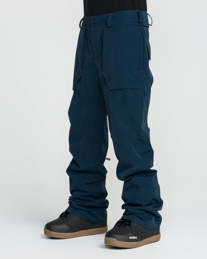 A person is shown from the waist down wearing Volcom Men’s Roan 20K Pants in dark blue cargo style, paired with black snow boots featuring brown soles, standing against a plain white background.