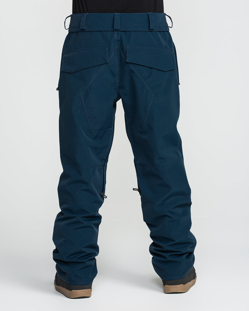 A person wearing Volcom Men’s Roan 20K Pants in dark blue and brown shoes, photographed from the back. The pants feature two large back pockets with flaps, reinforced stitching, and zippers near the lower legs.