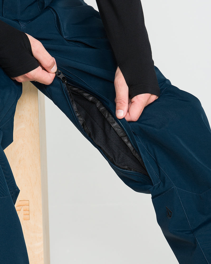 Wearing Volcom Men’s Roan 20K Pants in dark blue, a person sits on a wooden stool and unzips the side vent to reveal the mesh lining inside the waterproof pant leg.