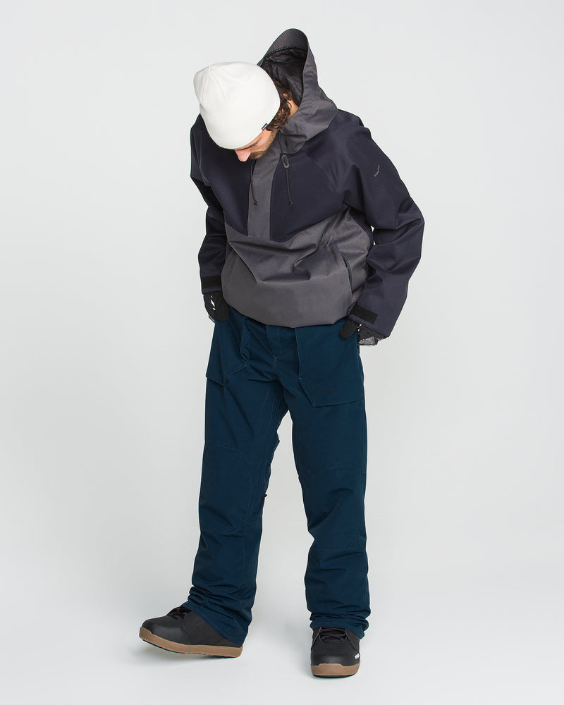 A person wearing a white beanie, dark jacket, and Volcom Men’s Roan 20K Pants in blue—crafted from recycled fabric for winter or snow activities—looks down at the ground against a plain white background.