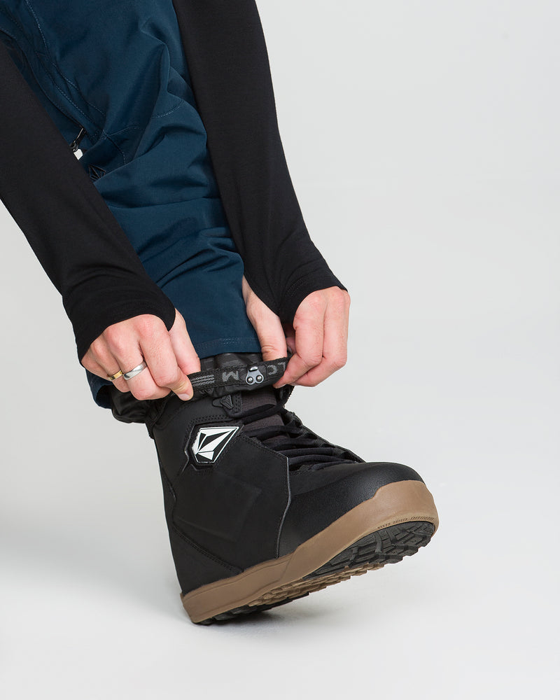 Wearing the Volcom Men’s Roan 20K Pants in blue, a person in a black long-sleeve top tightens the laces of a black snowboarding boot with a brown sole on their foot.