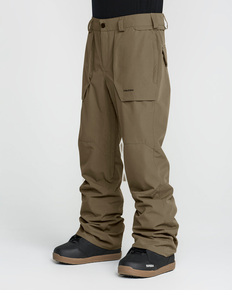 A person wearing Volcom Men’s Roan 20K Pants in olive green with black boots, standing against a plain white background. These pants are crafted from recycled fabric, feature convenient pockets, and display the VOLCOM logo on the upper thigh.